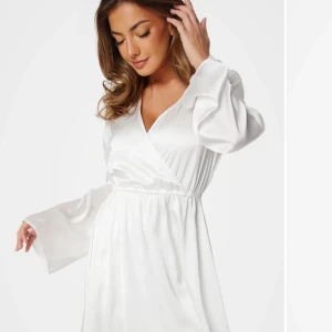 Vit omlottklänning - Säljer en elegant vit omlottklänning med långa ärmar och v-ringning. Klänningen har en mjuk och glansig yta som ger en lyxig känsla. Perfekt för speciella tillfällen eller student!! 💫Den är använd 1 gång (på min studentmottagning) så den är i mycket gott skick!!🌟