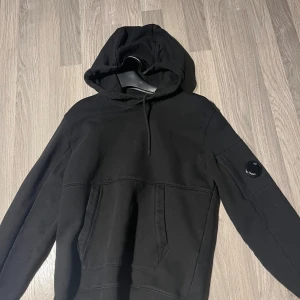 Svart hoodie från C.P. Company - Säljer en stilren svart hoodie från C.P. Company. Den har en klassisk design med huva och en ficka framtill. Storlek S 