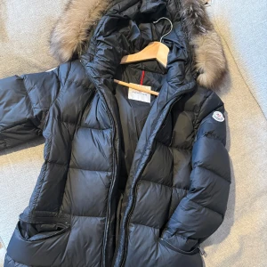 Svart dunjacka med pälsdetaljer från Moncler - Snygg svart dunjacka från Moncler med pälsdetaljer på huvan. Jackan har en lång design med dragkedja och två praktiska fickor framtill. Perfekt för kalla vår/vinterdagar. Som ny.          Skriv vid intresse!                                   