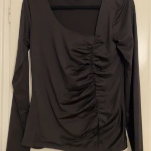 Svart långärmad topp med rynkdetalj - Snygg svart långärmad topp med rynkdetalj framtill. Perfekt för en stilren look. Tillverkad i ett mjukt och bekvämt material. Helt oanvänd. 