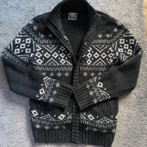 Stickad cardigan - Säljer en snygg mörkgrå stickad kofta med traditionellt mönster i vitt och grått. Koftan har knappar och dragkedja framtill samt ribbade muddar. Perfekt för kyliga dagar! Skick 10/10! Storlek M. Pris är ej hugget i sten så kom med pris förslag🤩!