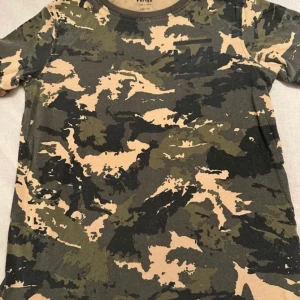 Camouflage t-shirt från WARP - Säljer en cool camouflage t-shirt från WARP. Den har ett klassiskt mönster i gröna och beige toner. Perfekt för en avslappnad stil.