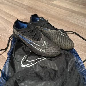 Nike phantom gx elite fg - Säljer ett par svarta Nike fotbollsskor med blå och vita detaljer. Skorna har snörning och en modern design med tydliga Nike-loggor. Perfekta för fotbollsplanen!