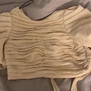 Beige bolerotopp med knyt - Charmig beige bolerotopp med knyt bak. Toppen har en texturerad yta och korta ärmar, vilket ger en unik look. Perfekt för att lägga till en touch av stil till din outfit.