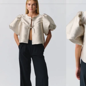 Beige kortärmad topp med puffärmar - Trendig beige topp med stora puffärmar och knytning framtill. Den har en unik design med korta ärmar och en öppen front som knyts ihop. Perfekt för att göra ett statement.