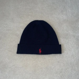 Mörkblå mössa med röd logga - Snygg mörkblå mössa från Ralph Lauren med en broderad röd logga framtill. Tillverkad av 100% ull, vilket gör den perfekt för kyliga dagar. Mössan har en klassisk design med uppvikt kant och är i storleken one size.