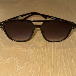 Svarta solglasögon - Snygga svarta solglasögon med en klassisk design och  tonade linser. Ramen har en stilren och enkel look med små silverdetaljer vid sidorna. Perfekta för en solig dag och ger en cool touch till din outfit.