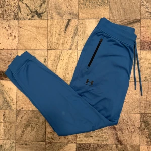 Blå mjukisbyxor från Under Armour - Säljer ett par blå mjukisbyxor från Under Armour i storlek M. Byxorna har en loose passform och är tillverkade i 100% polyester. De har snörning i midjan och dragkedjefickor på sidorna.