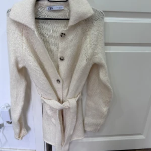 Beige stickad kofta från Zara - Mysig beige stickad kofta från Zara med knappar framtill och ett bälte i midjan. Perfekt för kyliga dagar med sin långa design och bekväma passform.