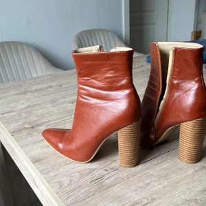 Bruna skinnstövletter med klack - Snygga bruna stövletter i skinn med en elegant spetsig tå och dragkedja på sidan. De har en robust klack som ger extra höjd och stil. Perfekta för att lyfta vilken outfit som helst.