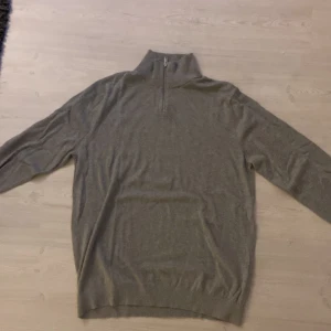 Grå tröja med dragkedja - Säljer en stilren grå tröja med dragkedja vid halsen. Tröjan har långa ärmar och ribbade kanter vid ärmslut och nederkant. Perfekt för kyligare dagar.