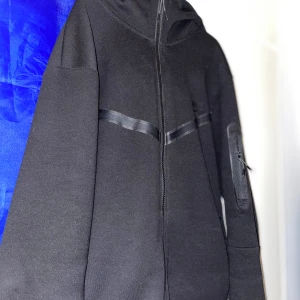 Svart kofta från Nike - Säljer en svart kofta från Nike med dragkedja och huva. Kofta har en diskret logga på bröstet och en praktisk ficka med dragkedja på ärmen. Perfekt för en sportig look. Notera att man inte kan öppna fickan på armen. Passar även storlek s.