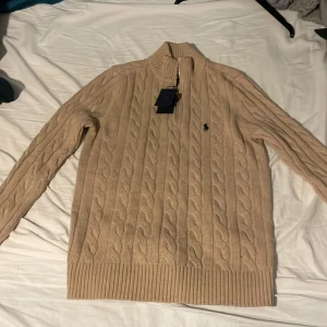 Beige stickad tröja från Polo Ralph Lauren - Säljer en snygg beige stickad tröja från Polo Ralph Lauren med dragkedja vid halsen. Tröjan har ett klassiskt flätmönster och ribbade muddar vid ärmslut och nederkant. Perfekt för kyligare dagar!