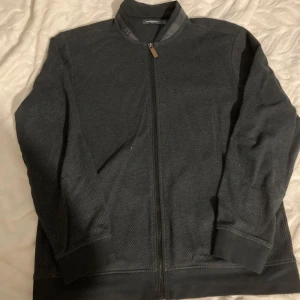 Grå kofta från Dressmann - Säljer en stilren grå kofta från Dressmann med dragkedja framtill. Koftan har en diskret textur och ribbade muddar vid ärmslut och nederkant. Perfekt för en avslappnad look.