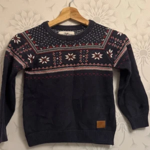 Mörkblå stickad tröja med mönster - Säljer en mysig mörkblå stickad tröja med ett traditionellt mönster i vitt och rött. Tröjan har långa ärmar och ribbstickade muddar vid ärmslut och nederkant. Perfekt för kyliga dagar!