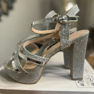 Glittriga silverfärgade sandaler med klack - Snygga sandaler i silver med glittrig yta och hög klack. De har flera remmar och en justerbar spänne runt ankeln för en säker passform. Perfekta för att lysa upp vilken outfit som helst.
