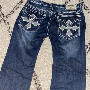Olimp jeans - Snygga blå jeans från Olimp med unika broderade korsdetaljer på bakfickorna. Jeansen har en bootcut-stil och är dekorerade med vita sömmar och röda knappar. Perfekta för dig som vill sticka ut!