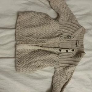 Beige stickad kofta från Little Pomp de Lux - Mysig beige stickad kofta från Little Pomp de Lux med ett fint flätmönster. Koftan har ribbade detaljer upptill och knappar framtill. Perfekt för kyliga dagar! Den kommer tvättas och strykas innan den skickas! ⭐️