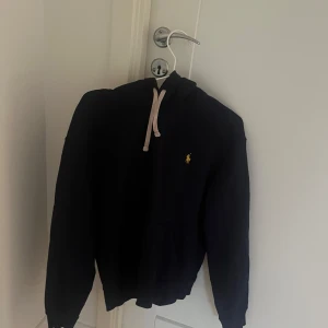Svart hoodie från Ralph Lauren - Snygg svart hoodie från Ralph Lauren med klassisk logga på bröstet. Den har en justerbar huva med vita snören och långa ärmar. Perfekt för en avslappnad stil.