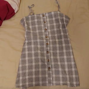 Grårutig klänning - Grårutig minidress med knytband och dragkedja där bak. Knappar framtill och adjusterbara band. Har en söm som blivigt lagad men den är så osynlig att jag glömt vart den är. Står storlek 10 alltså M, fungerar även på S.