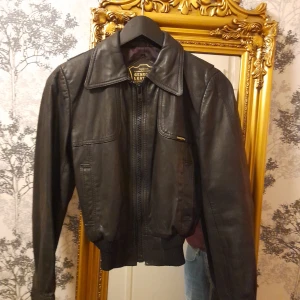 Vintage Svart skinnjacka från Petroff - Snygg svart skinnjacka från Petroff i äkta läder. Jackan har en klassisk krage och dragkedja framtill. Den är långärmad med ribbade muddar vid ärmslut och nederkant för en bekväm passform. Använd fåtal ggr på 60 talet. Märkt stl 14 år passar en vuxen stl 34/36