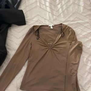 Beige långärmad topp med ringdetalj - Säljer en stilren beige långärmad topp med en dekorativ ringdetalj framtill. Perfekt för både vardag och festliga tillfällen. Toppen har en smickrande passform och är tillverkad i ett mjukt material.