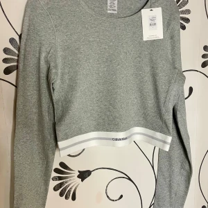 Grå ribbad tröja från Calvin Klein - Säljer en stilren grå ribbad tröja från Calvin Klein. Tröjan är croppad med långa ärmar och har en vit kant med logotypen vid nederkanten. Perfekt för en avslappnad look. Nypris 599:- prislapp kvar
