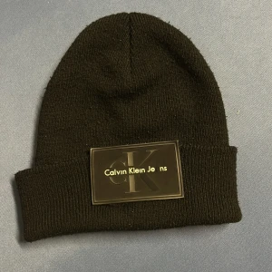 Svart mössa från Calvin Klein - Snygg svart mössa från Calvin Klein med ett stort märke framtill. Perfekt för att hålla värmen med stil. Den ribbade kanten ger en skön passform.