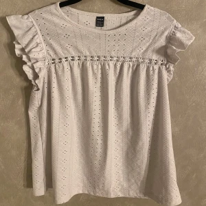 Vit blus från Shein - Säljer en söt vit blus från Shein med volangdetaljer på ärmarna och ett fint broderat mönster. Blusen är i 100% bomull och har en rund halsringning. Perfekt för en somrig look!🌞🥰