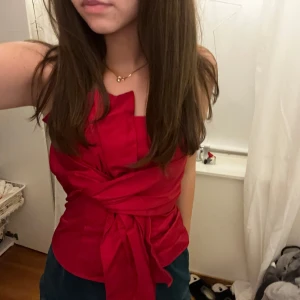 Röd axelbandslös topp med knyt - Snygg röd axelbandslös topp med knyt framtill. Perfekt för en kväll ute eller en speciell tillställning. Toppen har en elegant design som framhäver midjan.