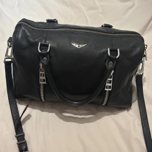 Zadig &Voltaire Sunny medium väska - Snygg svart skinnväska med silverfärgade detaljer och dubbla dragkedjor framtill. Väskan har både handtag och en justerbar axelrem för flexibel användning. Perfekt för att bära med sig det nödvändigaste med stil. Nypris: 5 550kr