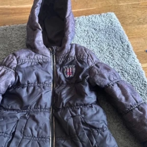 Grå pufferjacka med broderad detalj - Säljer en grå pufferjacka med huva och dragkedja. Jackan har en broderad detalj på bröstet med texten 'ATHL. CREW'. Perfekt för kyliga dagar.