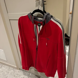 Röd hoodie och byxor från Boss - Snyggt set med röd hoodie och matchande byxor från Boss. Hoodien har dragkedja och snörning i huvan, medan byxorna har en bekväm passform med snörning i midjan. Båda plaggen har vita och grå ränder längs sidorna för en sportig look. Byxorna har storlek L men känns som M och hoodien har storlek M. 