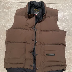 Brun dunväst från Canada Goose - Säljer en brun dunväst från Canada Goose med dragkedja och knappar. Västen har två stora fickor framtill och en hög krage för extra värme. Perfekt för kyliga dagar.