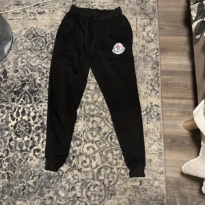 Svarta och vita mjukisbyxor med logga - Säljer två par mjukisbyxor i svart och vitt med en snygg logga på benet. Byxorna har snörning i midjan och muddar vid bensluten för en bekväm passform. Perfekta för en avslappnad stil. Pris kan diskureas man får båda för 100