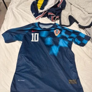 Blå fotbollströja från Nike - Säljer en blå fotbollströja från Nike med Kroatiens landslagsemblem och nummer 10 på framsidan. Tröjan har korta ärmar och ett schackrutigt mönster på axlarna. Namnet 'Modrić' och nummer 10 är tryckt på baksidan. Perfekt för fotbollsfans! Pris kan diskuteras 