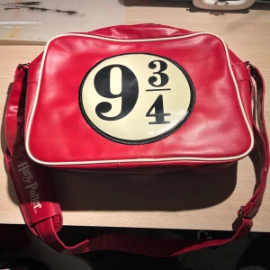 Röd axelväska med Harry Potter-tema - Röd axelväska med det ikoniska 9 3/4-märket från Harry Potter. Väskan har en justerbar axelrem, den är lite använd så har lite märken.