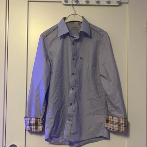 Ljusblå skjorta från Burberry - Snygg ljusblå skjorta från Burberry med klassiskt rutigt mönster på insidan av manschetterna. Skjortan har långa ärmar och knappar framtill. Perfekt för en stilren look.