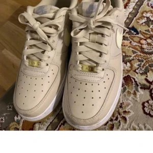 Beige Nike Air Force sneakers - Snygga beige Nike Air Force sneakers med klassisk design och perforerade detaljer för ventilation. Skorna har en vit sula och ikonisk Nike-logga på sidan. Perfekta för en stilren look.