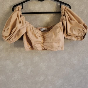 Beige offshoulder topp från Deezee - Trendig beige offshoulder topp från Deezee med puffiga ärmar och rynkdetaljer. Perfekt för en stilfull look.