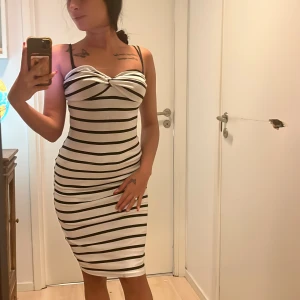 Randig klänning med smala axelband - Snygg och figurnära klänning med svartvita ränder utan axelband ( har bh på bilden ) . Klänningen har en vacker twistdetalj vid bysten och är knälång, perfekt för en stilren look.