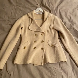 Beige kofta  från Chiara Forthi - Elegant beige kavaj från Chiara Forthi med dubbelknäppning och gyllene knappar. Den har en stilren krage och långa ärmar, perfekt för en chic look. Materialet är stickat vilket ger en mjuk och bekväm känsla.