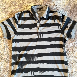 Randig pikétröja från J.Lindeberg - Snygg pikétröja från J.Lindeberg med ett unikt svart och grått randigt mönster. Tröjan har en klassisk krage med knappar och korta ärmar. Perfekt för en stilren och avslappnad look.