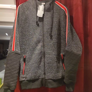 Svart och grå jacka med röda detaljer från CINC - Säljer en snygg svart och grå jacka från CINC med röda och vita ränder längs ärmarna. Jackan har dragkedja och ribbade detaljer på ärmarna. Perfekt för en cool och avslappnad stil.