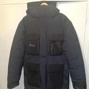 Svart Gore-Tex jacka från Sail Racing - Säljer en nästintill oanvänd svart Gore-Tex jacka från Sail Racing. Jackan har flera praktiska fickor med dragkedjor och en quiltad insida för extra värme. Perfekt för kalla och blöta dagar med sin vattentäta och vindtäta design. Den har en hög krage och justerbar huva. Säljer pga för stor i storleken.