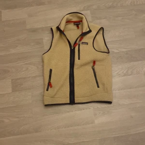 Beige fleeceväst från Patagonia - Säljer en beige fleeceväst från Patagonia med dragkedja och flera praktiska fickor. Västen har svarta detaljer och en liten logga på bröstet. Perfekt för lager-på-lager under kyliga dagar.