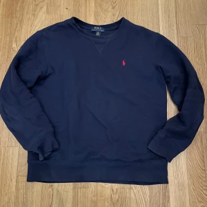 Ralph lauren tröja - Säljer en klassisk mörkblå tröja från Ralph Lauren med det ikoniska röda logotypbroderiet på bröstet. Tröjan har långa ärmar och en rund halsringning. Perfekt för en stilren och avslappnad look. Storlek är 14-16 år som motsvara XS.