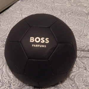 Svart fotboll från Boss Parfums - En stilren svart fotboll med Boss Parfums logotyp i vitt. Perfekt för den som älskar både sport och stil. Bollen har en klassisk design med pentagonmönster och är tillverkad i ett slitstarkt material.