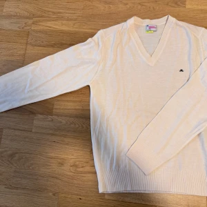 Vit tröja från J.Lindeberg - Säljer en stilren beige tröja från J.Lindeberg med v-ringning. Tröjan har långa ärmar och ribbade muddar vid ärmslut och nederkant. Perfekt för en klassisk look.