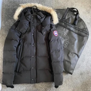 Svart dunjacka från Canada Goose - Säljer en svart dunjacka från Canada Goose med pälsdetalj på huvan. Jackan har en broderad logotyp på ärmen och stängs med dragkedja och knappar. Perfekt för kalla vinterdagar.